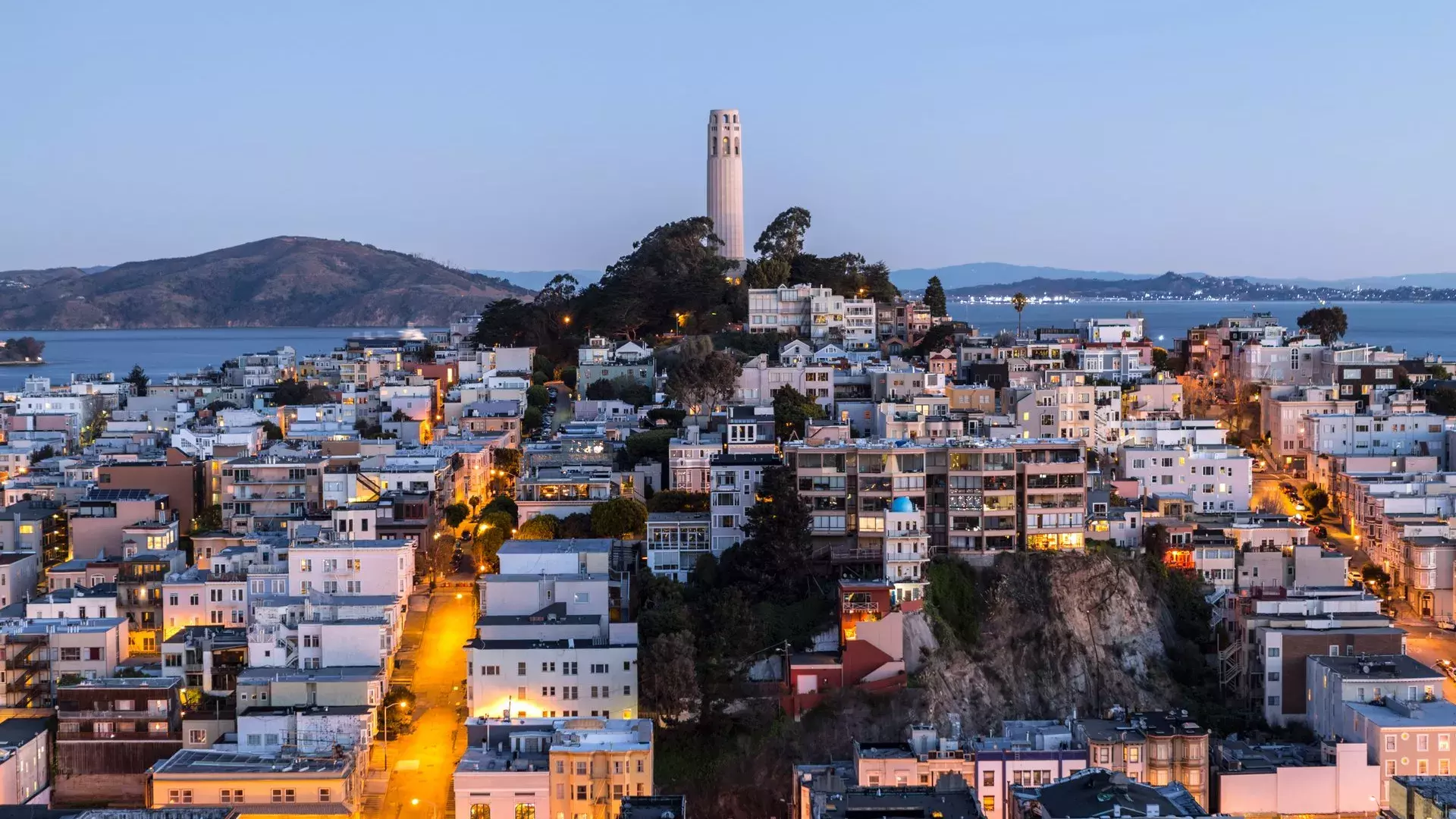 San Francisco's Coit Tower at dusk, with lighted streets before it and the San Francisco Bay behind it.