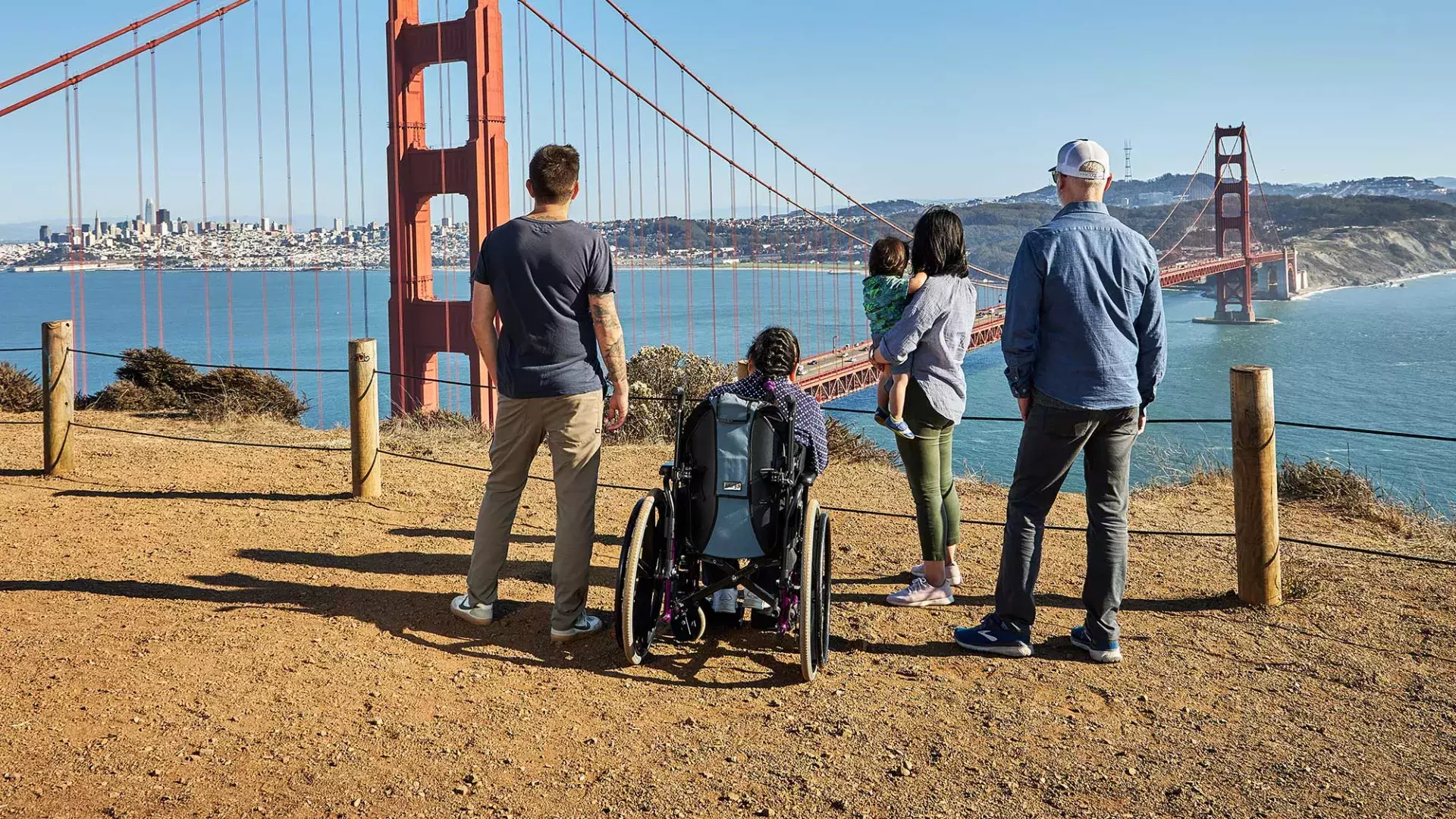 A group of people, including one person in a wheelchair, is seen from behind as they look at the Golden Gate Bridge from the Marin Headlands.