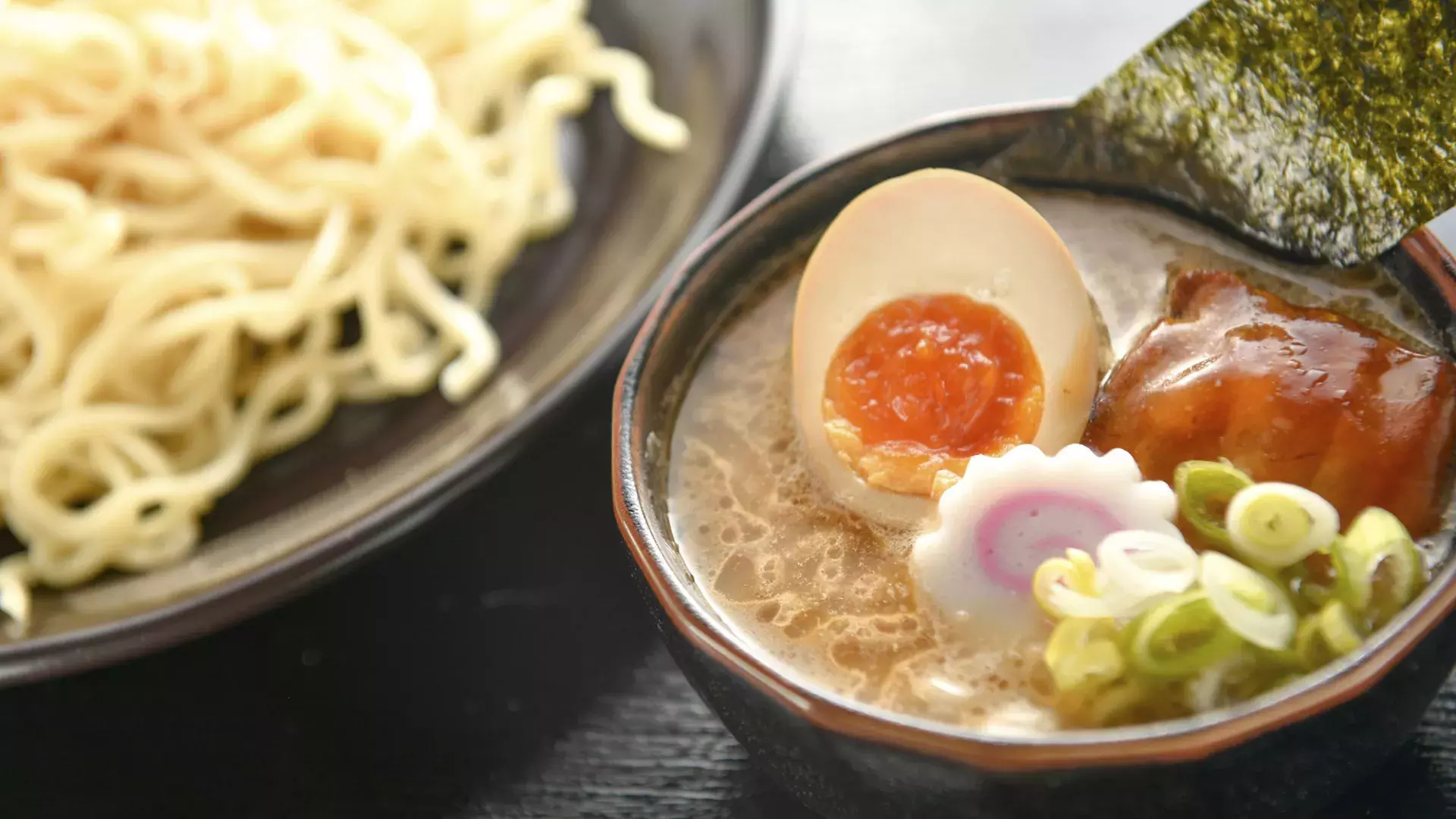 Close up shot of a bowl of noodles and a bowl of ramen soup with a poached egg, sliced in half.