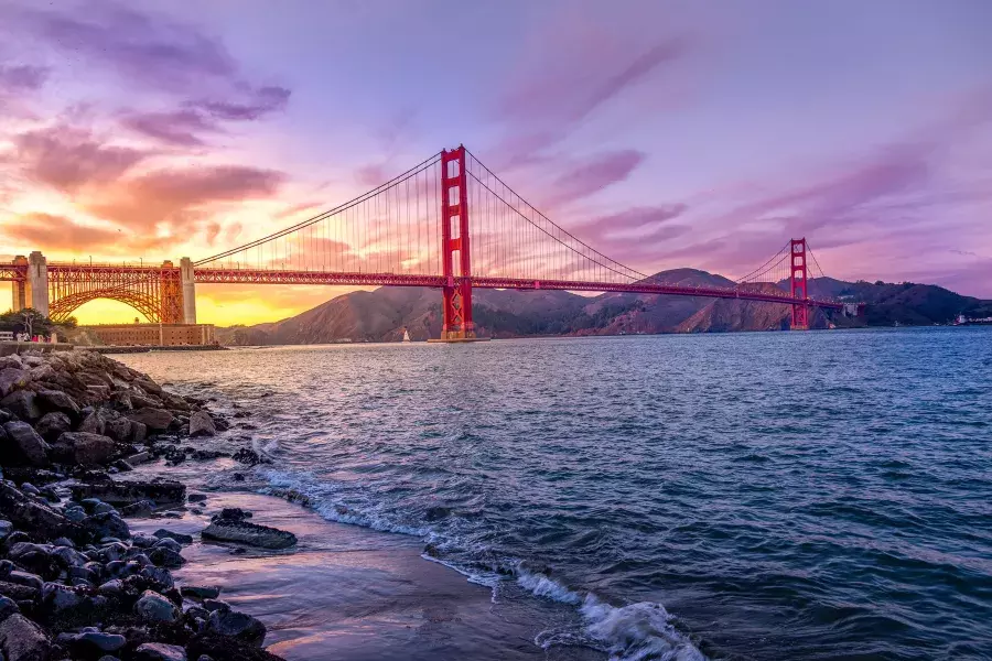 The Golden Gate Bridge at sunset with a multicolored sky and the San Francisco Bay in the foreground.