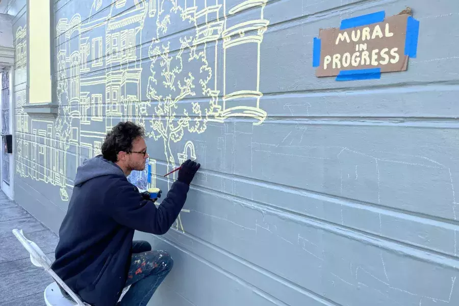 An artist paints a mural on the side of a building in the Mission District, with a sign taped onto the building that reads "Mural in Progress." San Francisco, CA.