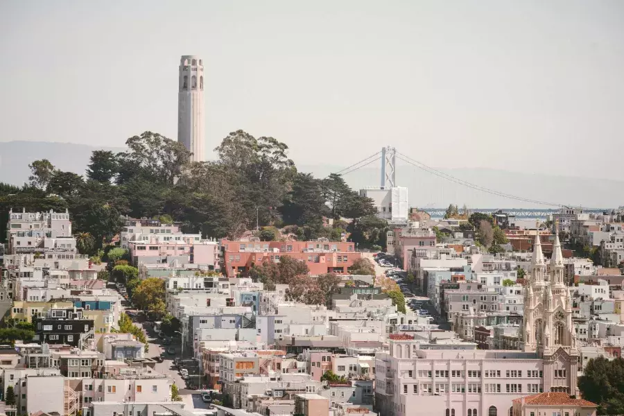 San Francisco's Coit Tower is pictured with the Bay Bridge in the background and a hill covered in houses in the foreground.