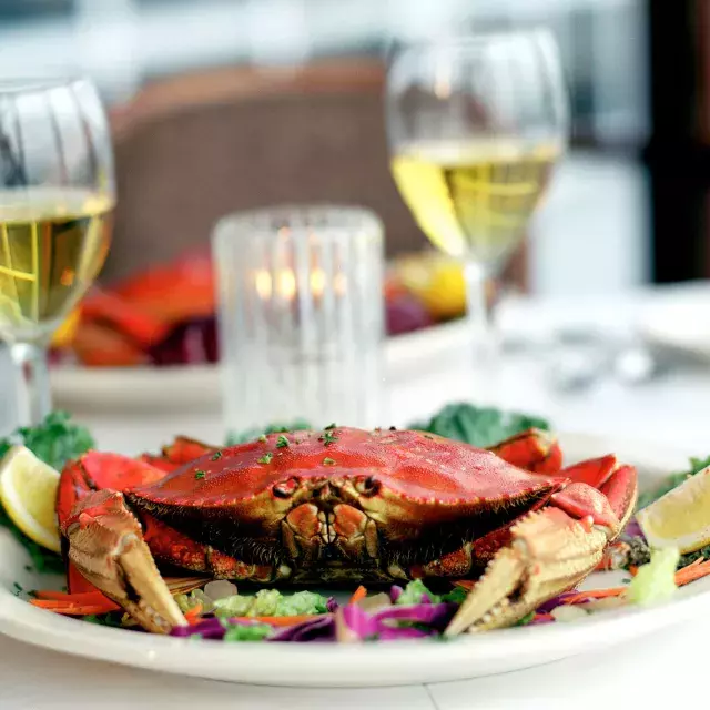 A Dungeness crab sits on a plate in a restaurant with two glasses of white wine in the background.