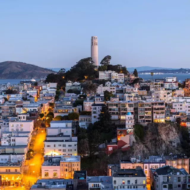 La Coit Tower de San Francisco au crépuscule, avec des rues éclairées devant elle et la baie de San Francisco derrière elle.