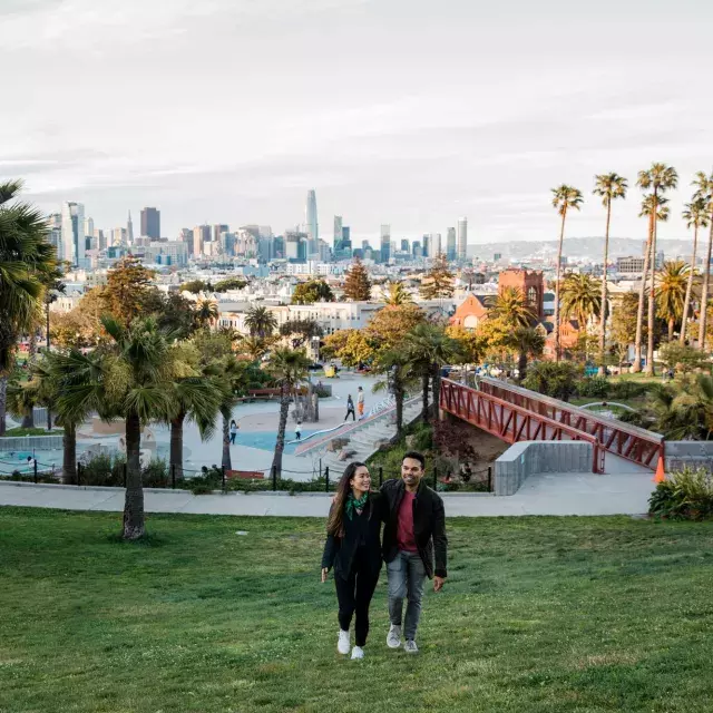 A couple walks toward the camera with Dolores Park and the San Francisco Skyline behind them.